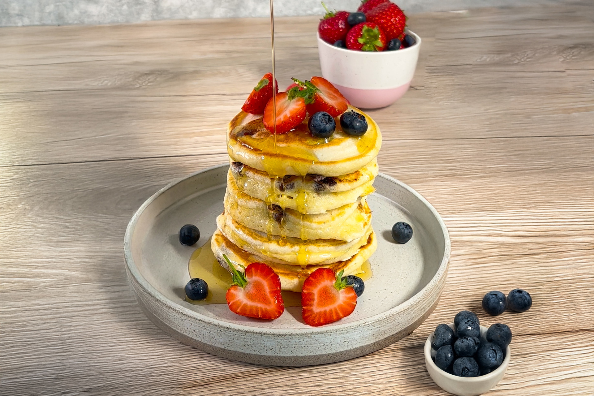 Auf einem Teller wurde ein Stapel frischer American Pancakes angerichtet. Die Pancakes sind mit frischen Beeren garniert und werden mit Ahornsirup übergossen.