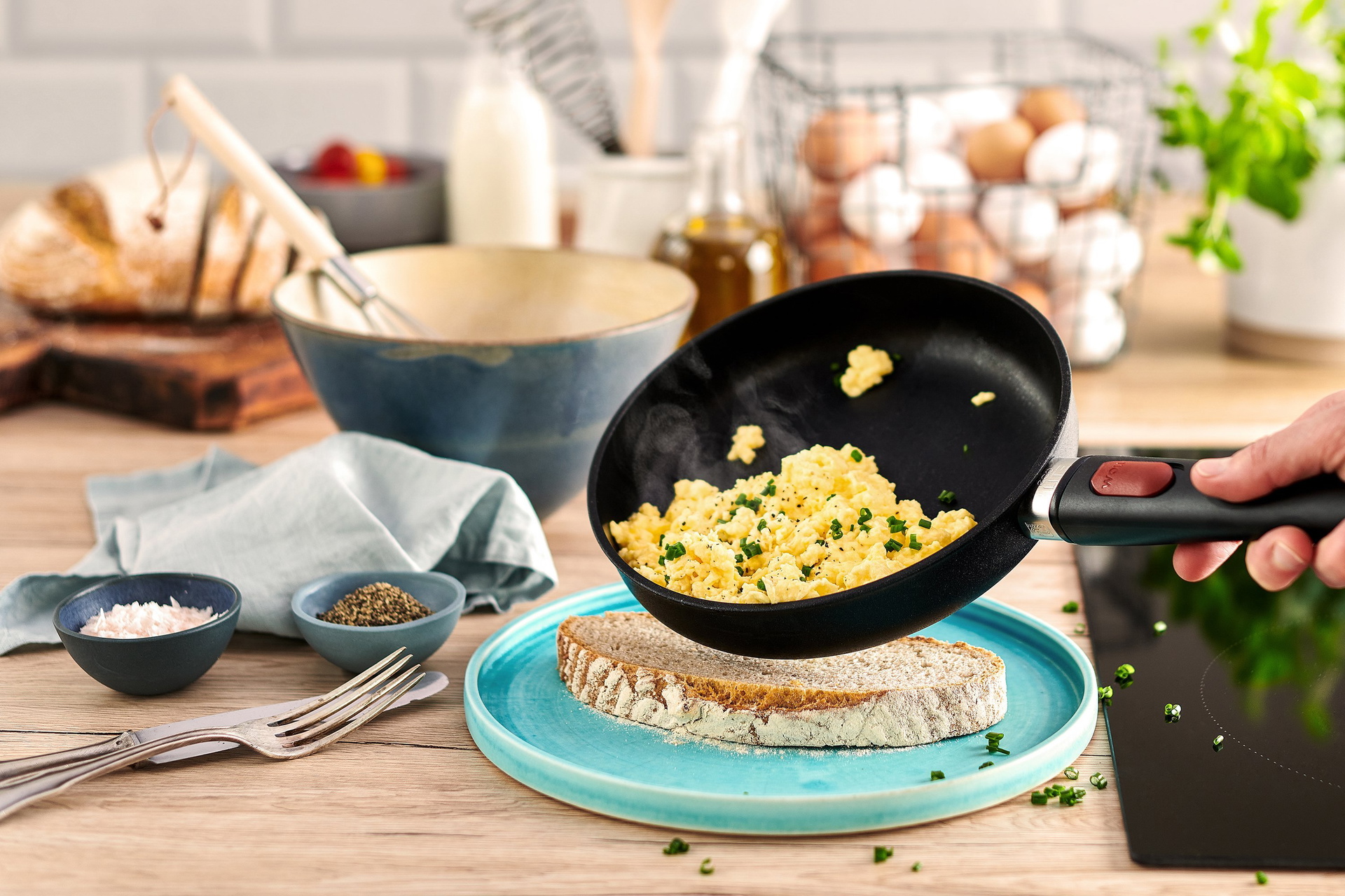 Aus einer runden, heißen Eco-Lite-Pfanne mit abnehmaren Stiel wird dampfendes Rührei direkt auf eine Scheibe Brot gegeben. Daneben stehen eine Schüssel, Besteck, Gewürze sowie mehrere Zutaten.