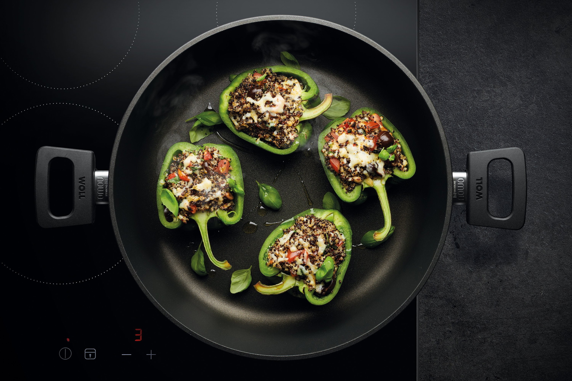 Auf dem Herd steht eine runde, heiße Kasserolle der Serie „Nowo” mit abnehmbarem Stiel. Darin befindet sich frisch zubereitete, gebackene grüne Paprika mit mediterranem Quinoa.