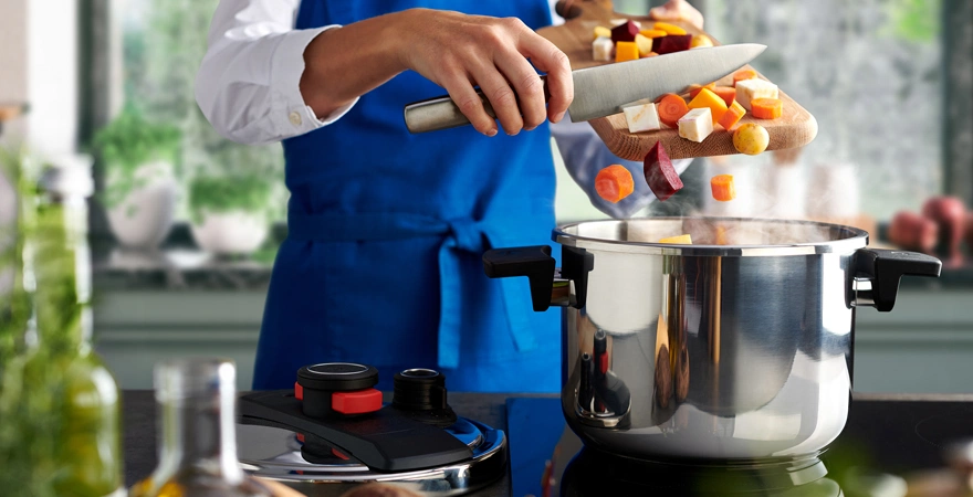 Auf dem Herd steht ein dampfender Pressure-Pro-Topf, während das frisch geschnittene Gemüse mit dem Messer vom Brett in den Topf gegeben wird. Der Deckel liegt links auf der Arbeitsplatte.