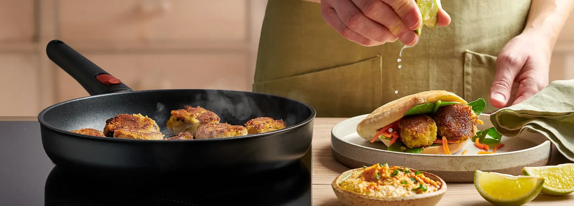 In einer runden, heißen Eco Lite QXR Pfanne werden frische Falafel angebraten. Auf dem Teller daneben liegt ein fertiges Fladenbrot gefüllt mit Falafel und eine Schüssel Hummus steht vor dem Teller.