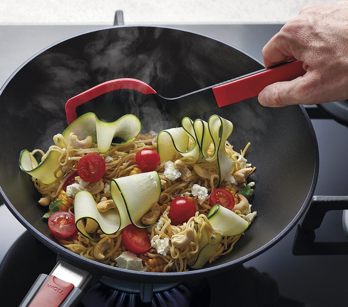 In einer runden Diamond Lite Wok- und Rührpfanne wird ein Nudelgericht zubereitet, das mit einer roten Cock it Pfannenschaufel umgerührt wird. 