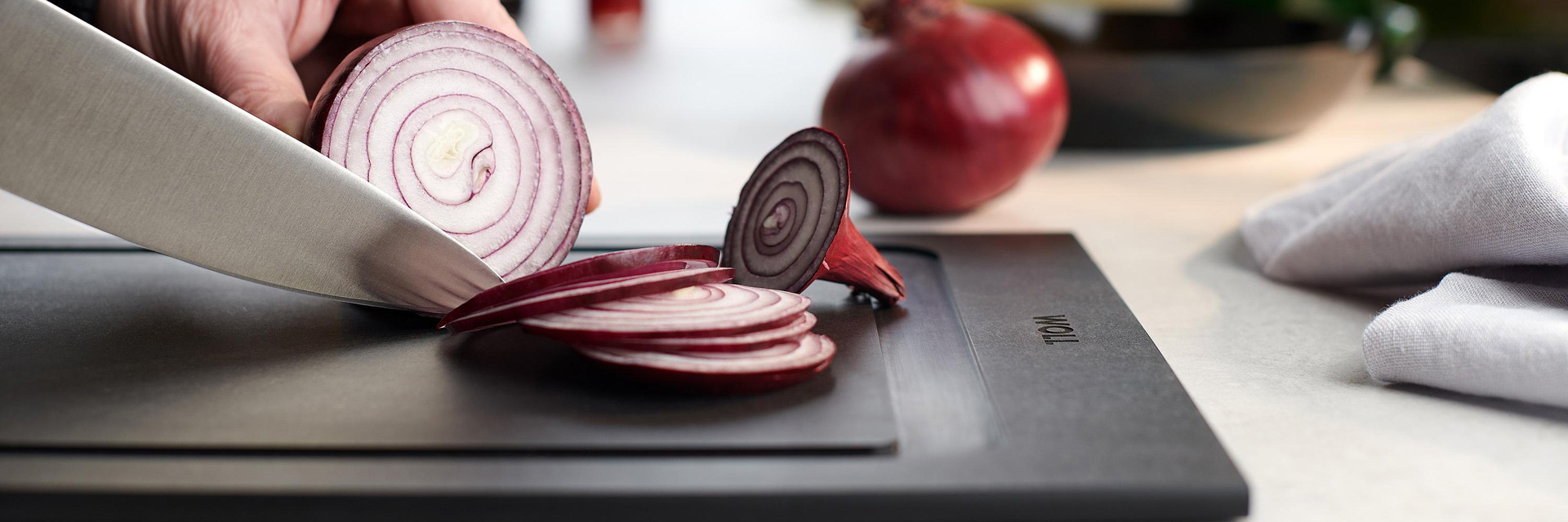 Auf einem Schneidebrett der Serie Cut & Serve werden mithilfe eines Messers Zwiebeln in Scheiben geschnitten. Im Hintergrund ist eine ganze Zwiebel zu sehen, seitlich liegt ein Küchentuch.