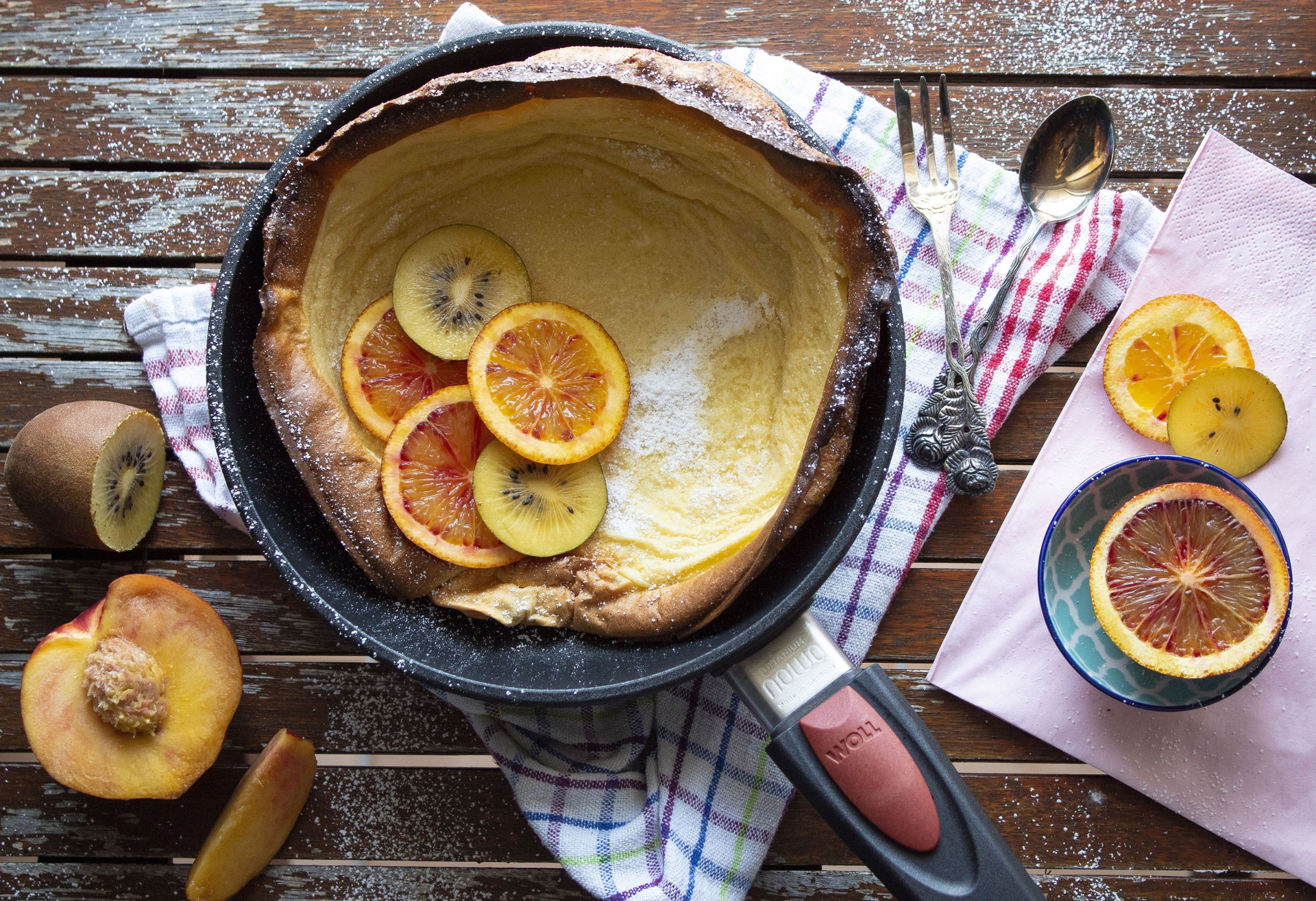 In einer runden, heißen Pfanne der Serie Nowo mit abnehmbarem Stiel wurde das Dutch Baby zubereitet. Er wurde mit Puderzucker bestreut und mit Kiwi-Scheiben und weiteren Obstscheiben dekoriert. Daneben liegt noch mehr Obst.