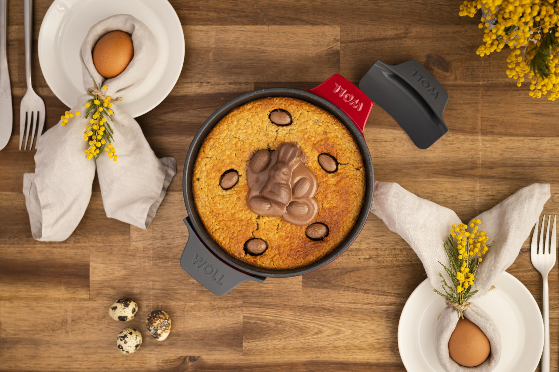 In einer runden Servierpfanne der Serie Iron befindet sich ein frisch gebackener Kuchen mit Schokoladen. Zur Sicherheit wurden Silikongriffe um die Griffe gelegt. Ein Griff liegt daneben, damit die Farbe erkennbar ist. Daneben liegen gekochte Eier auf Tel