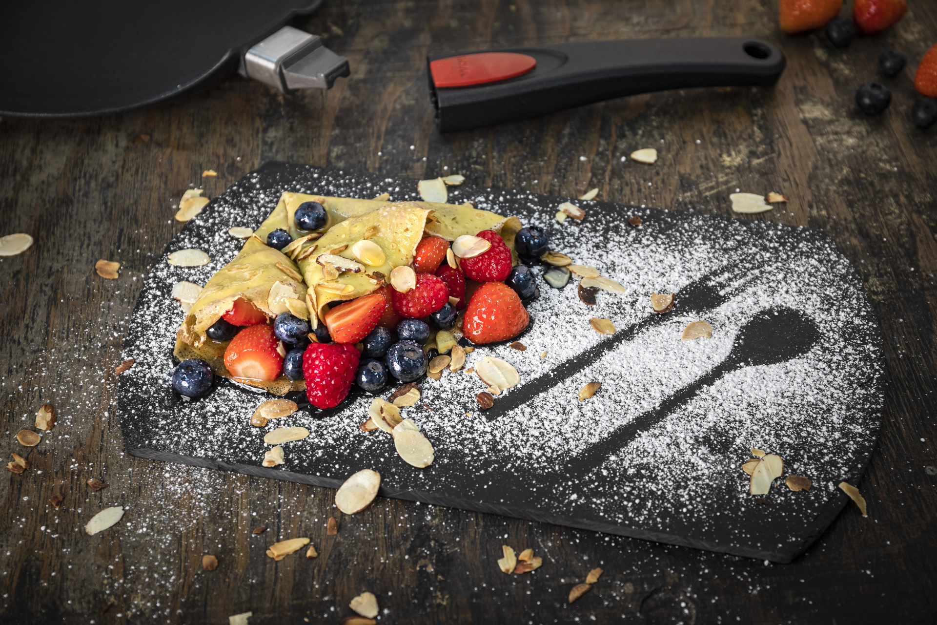 Titan Best Crêpepfannen-Set, 3-tlg., Ø 26 cm Auf einem Brett liegt ein mit Puderzucker bestäubter Crêpe. Dahinter befindet sich die Titan Best Crêpepfanne.