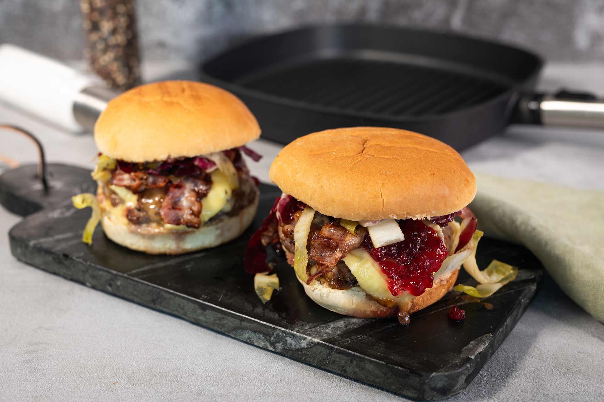 Auf einem Schneidebrett liegen frisch zubereitete Camembert Beef Burger. Dahinter steht eine viereckige Grillpfanne aus der Serie Logic mit abnehmbarem Stiel.