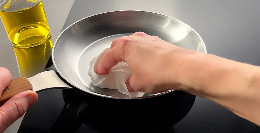 Auf dem Herd steht eine Carbon-Steel-Pfanne. In dieser wird mithilfe einer Küchenrolle Öl verteilt. Zur Fixierung wird die Pfanne am Griff festgehalten. Daneben steht eine Flasche mit neutralem Öl.