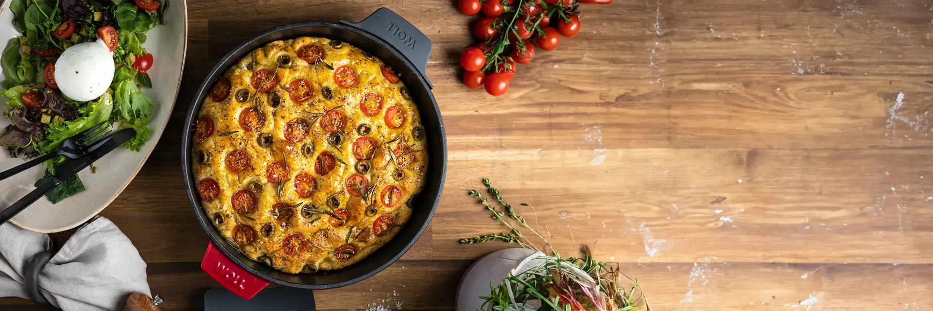 In der runden Kasserolle der Serie „Iron” befindet sich ein frisch gebackenes Focaccia, das mit Rosmarin und Meersalz garniert ist. Daneben stehen ein frischer Salat mit Burrata, frischen Kirschtomaten, ein Küchentuch, eine Mühle und frische Kräuter im To
