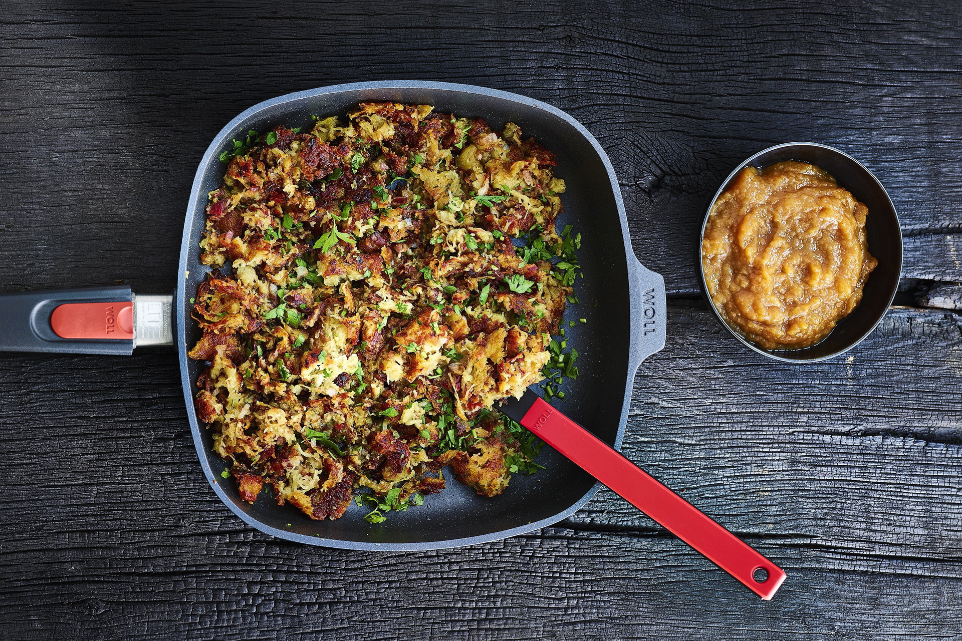 In einer viereckigen, heißen Diamond-Lite-Pfanne mit abnehmbarem Stiel befindet sich frisch zubereitetes Dippelabbes. In der Pfanne ist auch ein Küchenhelfer der Serie Cock it zu sehen. Daneben steht eine Schüssel mit Apfelmus.