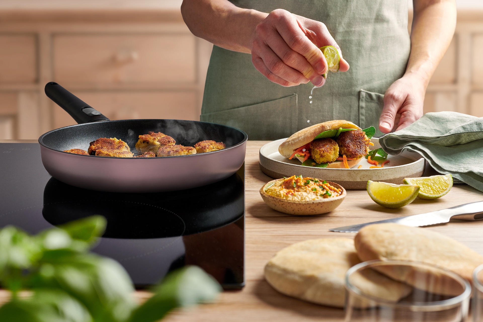 In einer runden, hellrosa Ecolour Pfanne werden Falafel angebraten. Ein gefülltes Fladenbrot liegt auf einem Teller. 