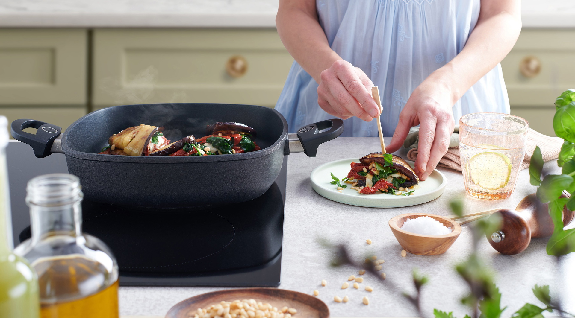 In einer viereckigen Pfanne der Serie Titan Best befinden sich dampfende Auberginen-Wraps. Auf einem Teller daneben wird ein frisch gebratener Wrap mit einem Holzspieß zusammengesteckt. Daneben stehen ein Glas Wasser und weitere Zutaten.