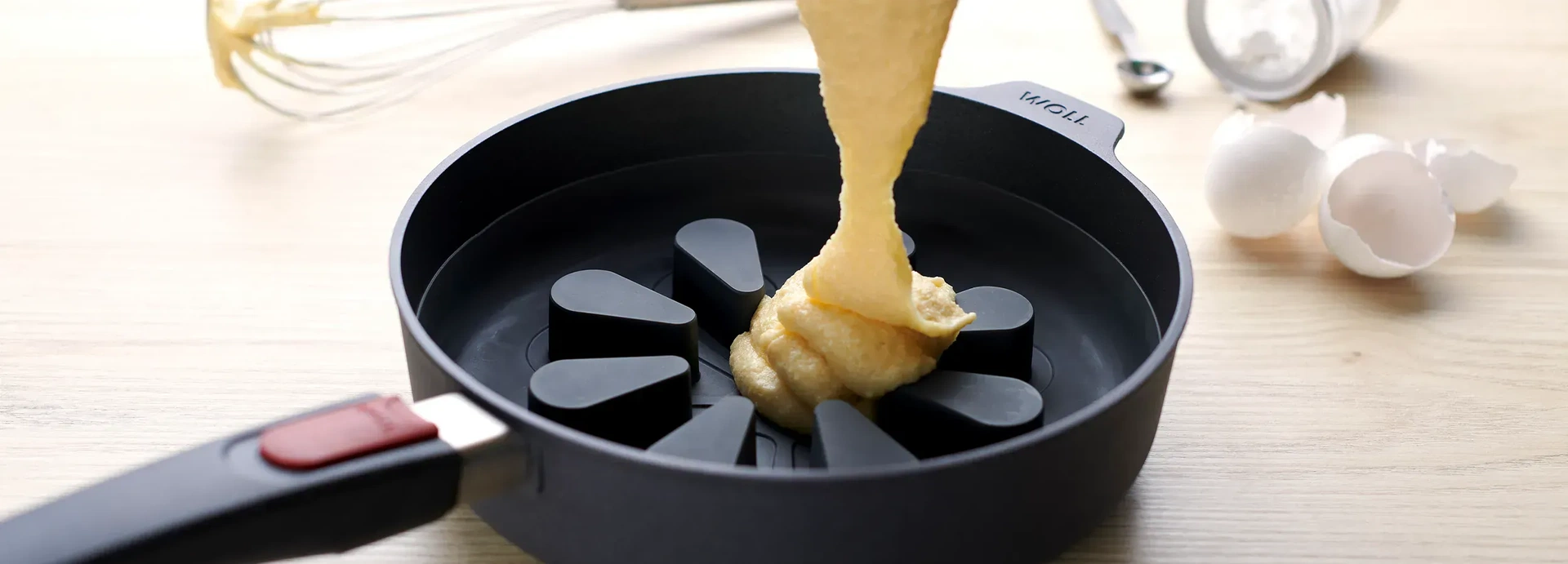 In eine Hochrandpfanne der Serie „Diamond Lite” mit einem Silikon-Backeinsatz der Serie „Bake it” wird heller Teig gefüllt. Im Hintergrund sind ein Schneebesen, ein kleiner Löffel, ein umgefallenes Glas mit Inhalt sowie Eierschalen zu sehen.