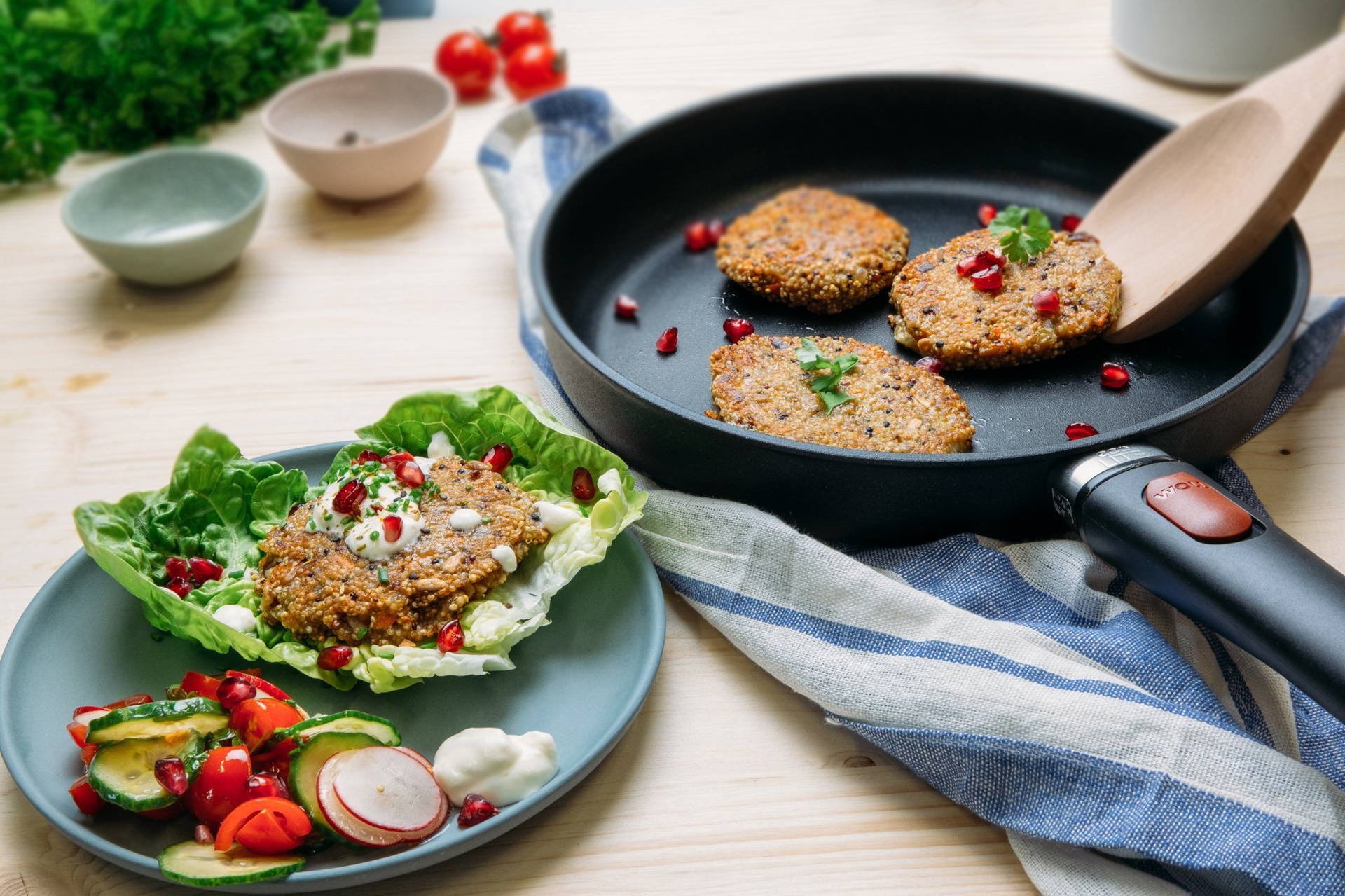 In einer runden Eco-Lite-QXR-Pfanne mit abgenommenem Stiel befinden sich frisch angebratene Quinoa-Bratlinge. Auf einem Teller daneben liegt ein weiterer Bratling auf einem Salatblatt.