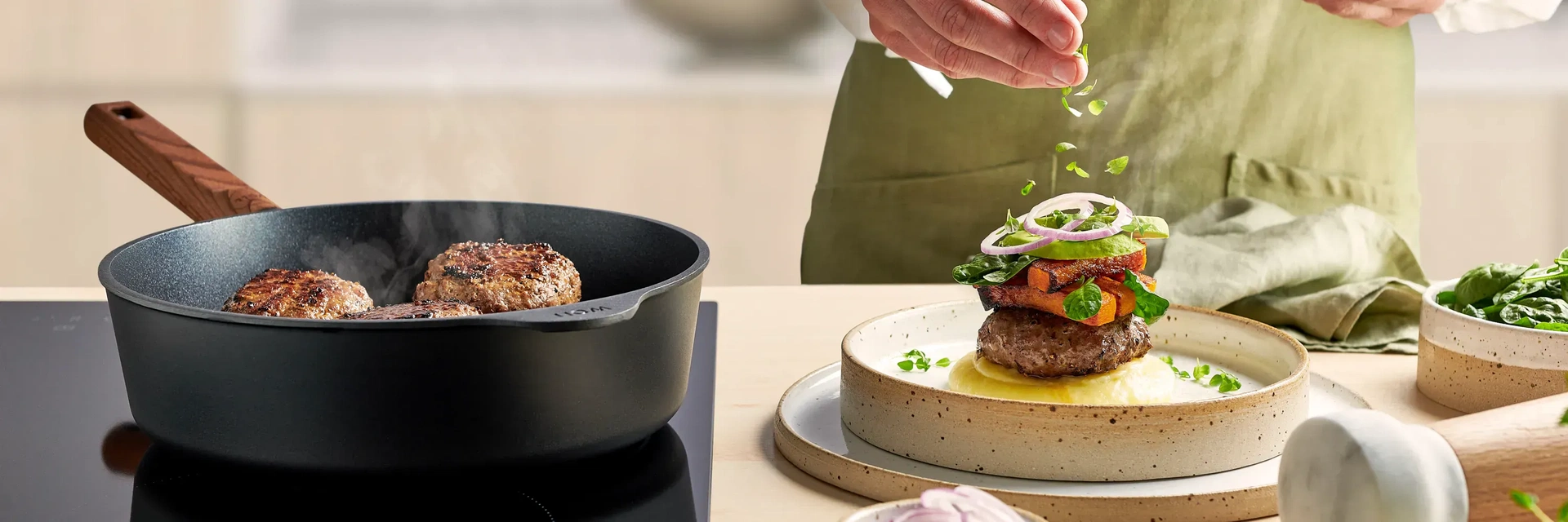 In einer runden Hochrandpfanne mit abgenehmbaren Griff der Serie Eco Logic QXR liegt fertig angebrachtes Fleisch. Daneben steht ein Teller mit Kartoffelpüree, darauf ein Stück Fleisch, garniert mit angebraten Karotten und etwas Salat.