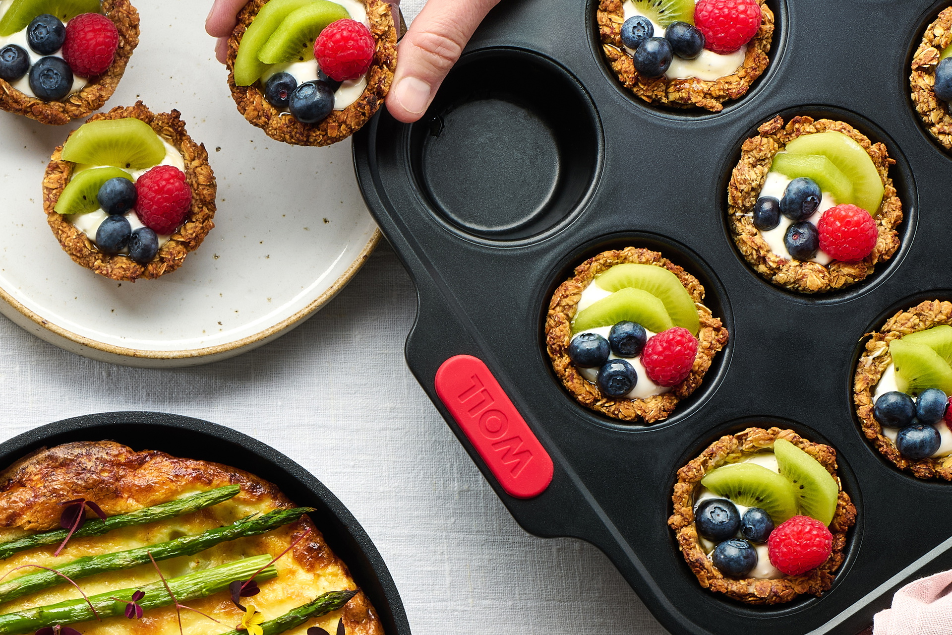 In einer runden Pfanne der Serie Eco Logic QXR befindet sich ein frisches Kartoffel-Spargel-Omelet. Der Stiel der Pfanne wurde abgenommen, damit sie auf den Tisch gestellt werden kann. Daneben wurden Granola-Cups aus der Muffinform auf den Teller gegebe