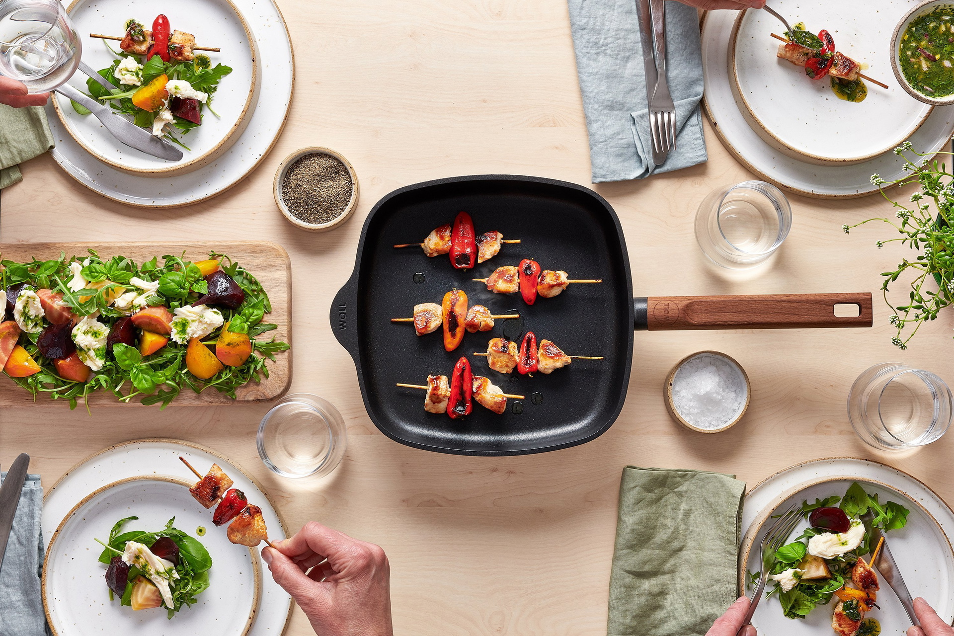 Auf dem Tisch steht eine viereckige Pfanne aus der Serie Eco Logic QXR mit abnehmbarem Stiel, in der sich frisch angebratene Fleischspieße befinden. Drumherum stehen Teller, Gläser, Gewürze, eine Pflanze und ein Salat, der auf einem Brett serviert wird.