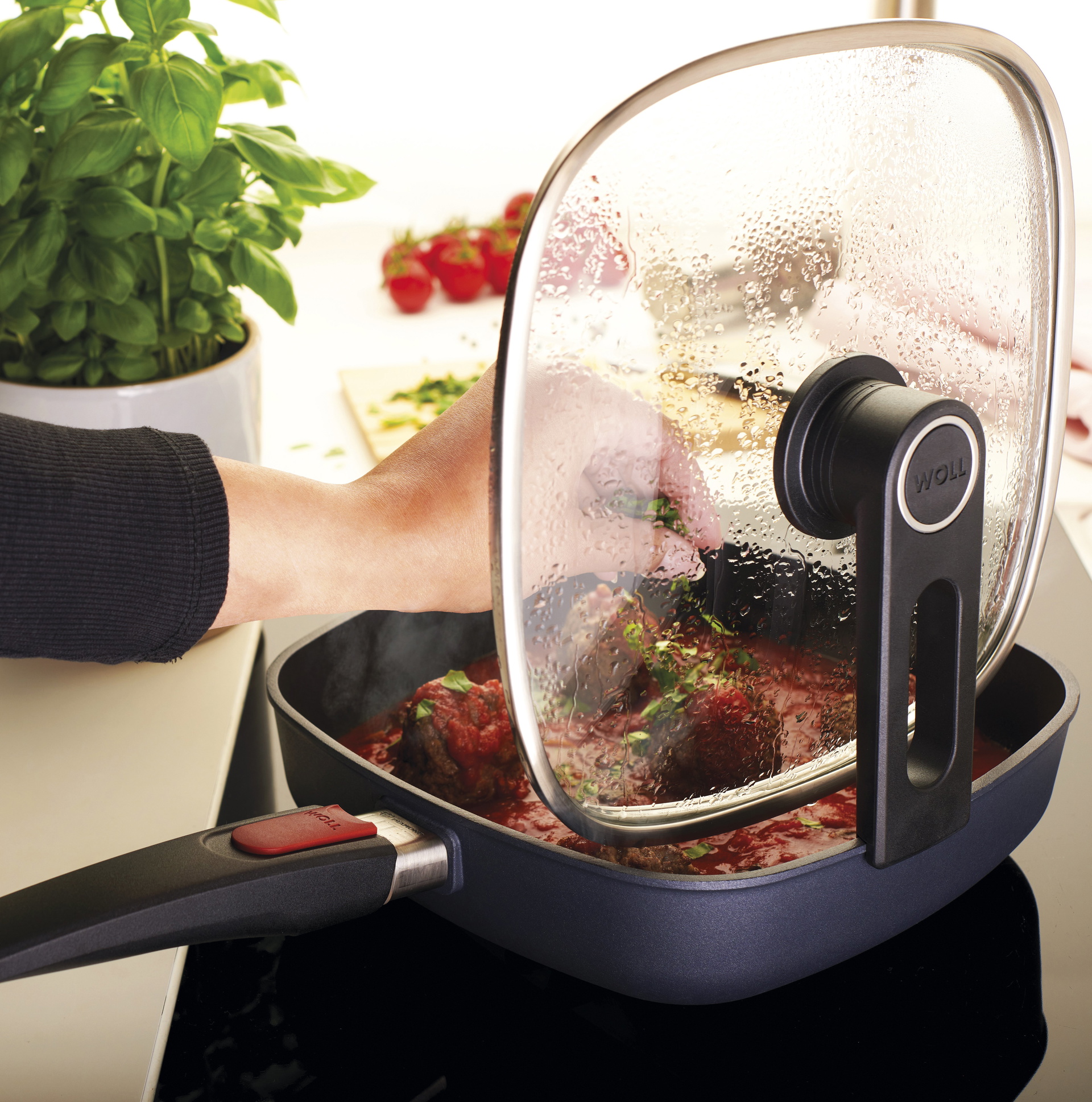 Smart-Lid_RGB In einer viereckigen, heißen Diamond-Lite-Pfanne befinden sich Fleischbällchen in Tomatensoße. Der Glasdeckel ist mit der verlängerten Griffseite der Smart-Lid-Serie an der Kante der Pfanne eingehängt. Im Hintergrund sind Kräuter in einem Topf, Kräuter au