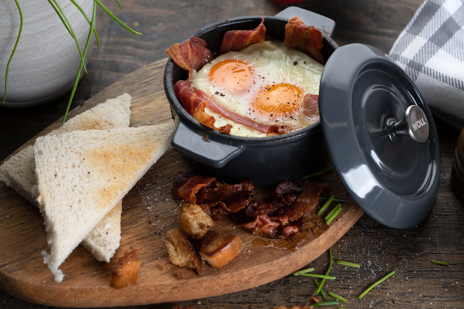 In einer heißen, runden Mini-Kasserolle aus der Serie „Iron” werden frisch gebackene Frühstücksmuffins mit Bacon und Ei serviert. Die Kasserolle steht auf einem Brett mit frischen Toastscheiben und Bacon.