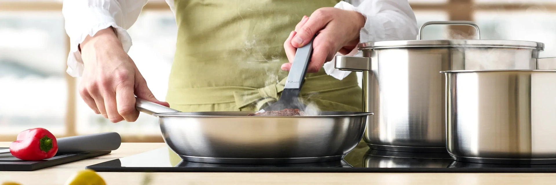 Auf dem Herd stehen eine runde Pfanne und Töpfe der Serie „Steel”. In der Pfanne wird Fleisch angebraten, das mit einem Pfannenwender gewendet wird. Daneben liegt ein Schneidebrett mit einem Messer und einer Paprika darauf.