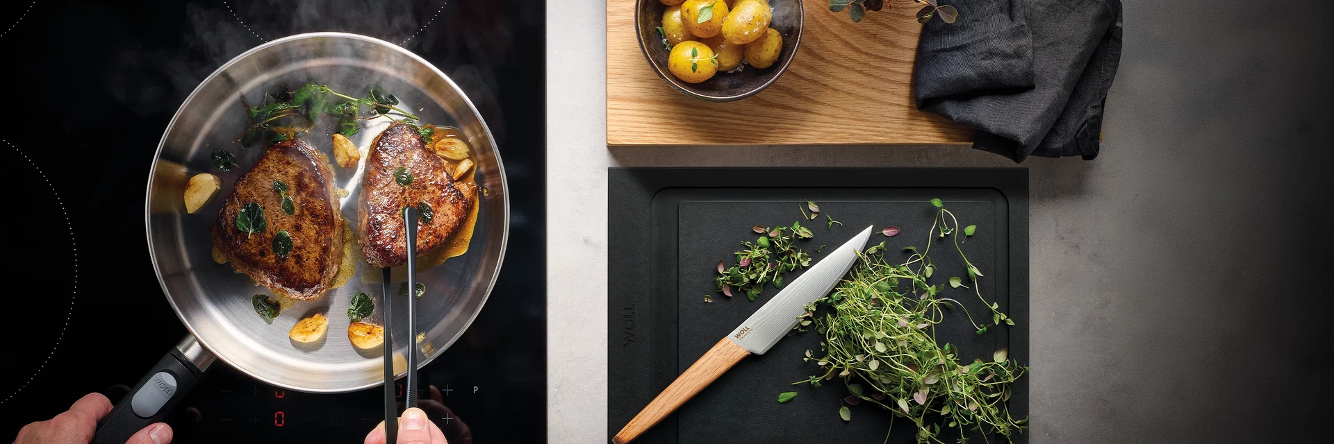 In einer heißen, runden Logic-Steel-Pfanne werden zwei Fleischstücke angebraten. Daneben liegt ein Schneidebrett mit frischen Kräutern und einem Messer darauf. In der Nähe steht eine Schüssel mit gekochten Kartoffeln.