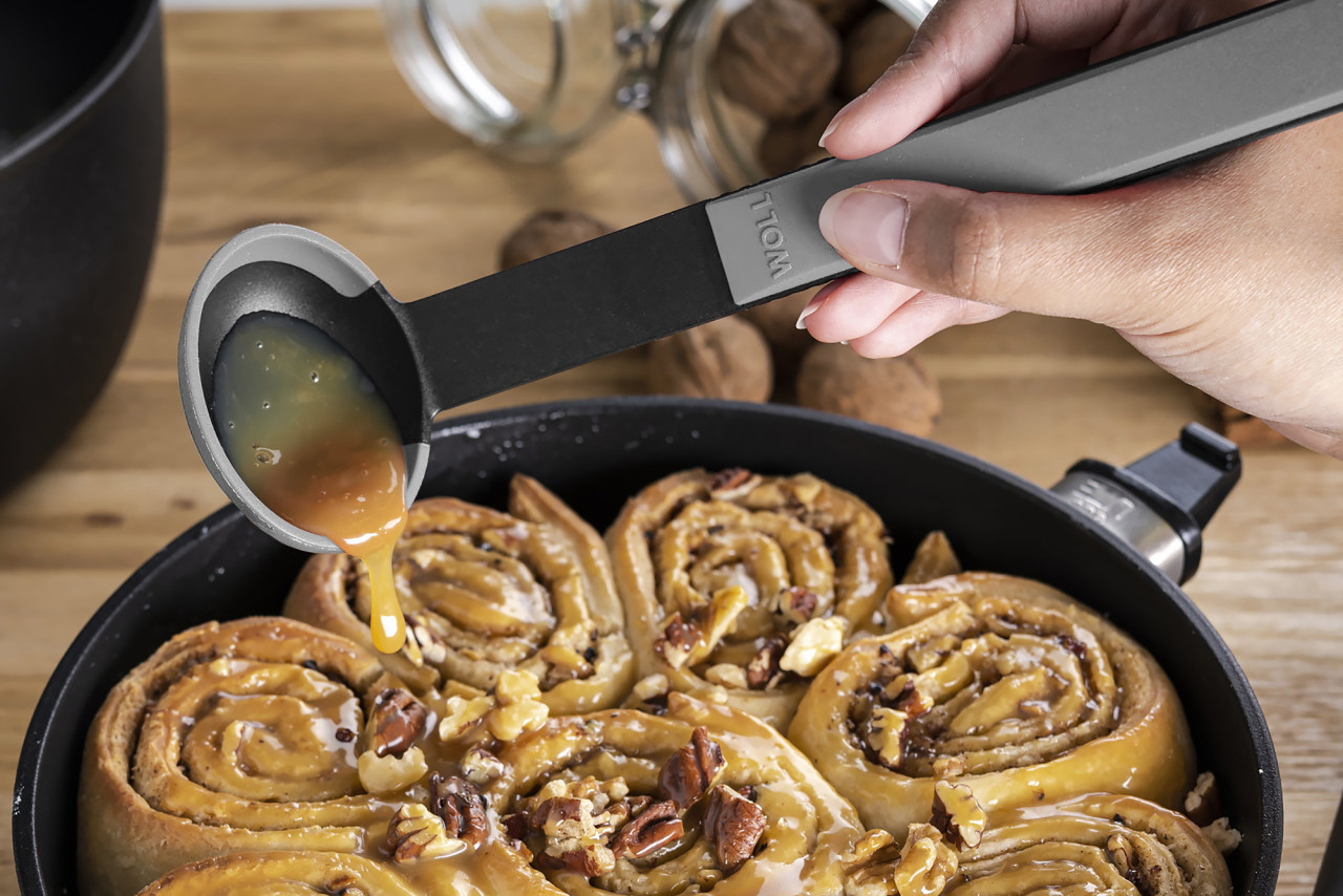 In einer runden Eco-Lite-QXR-Pfanne befinden sich Zimtschnecken, diese werden mit einem grauen Cock it Saucenlöffel mit Karamellsoße übergossen.