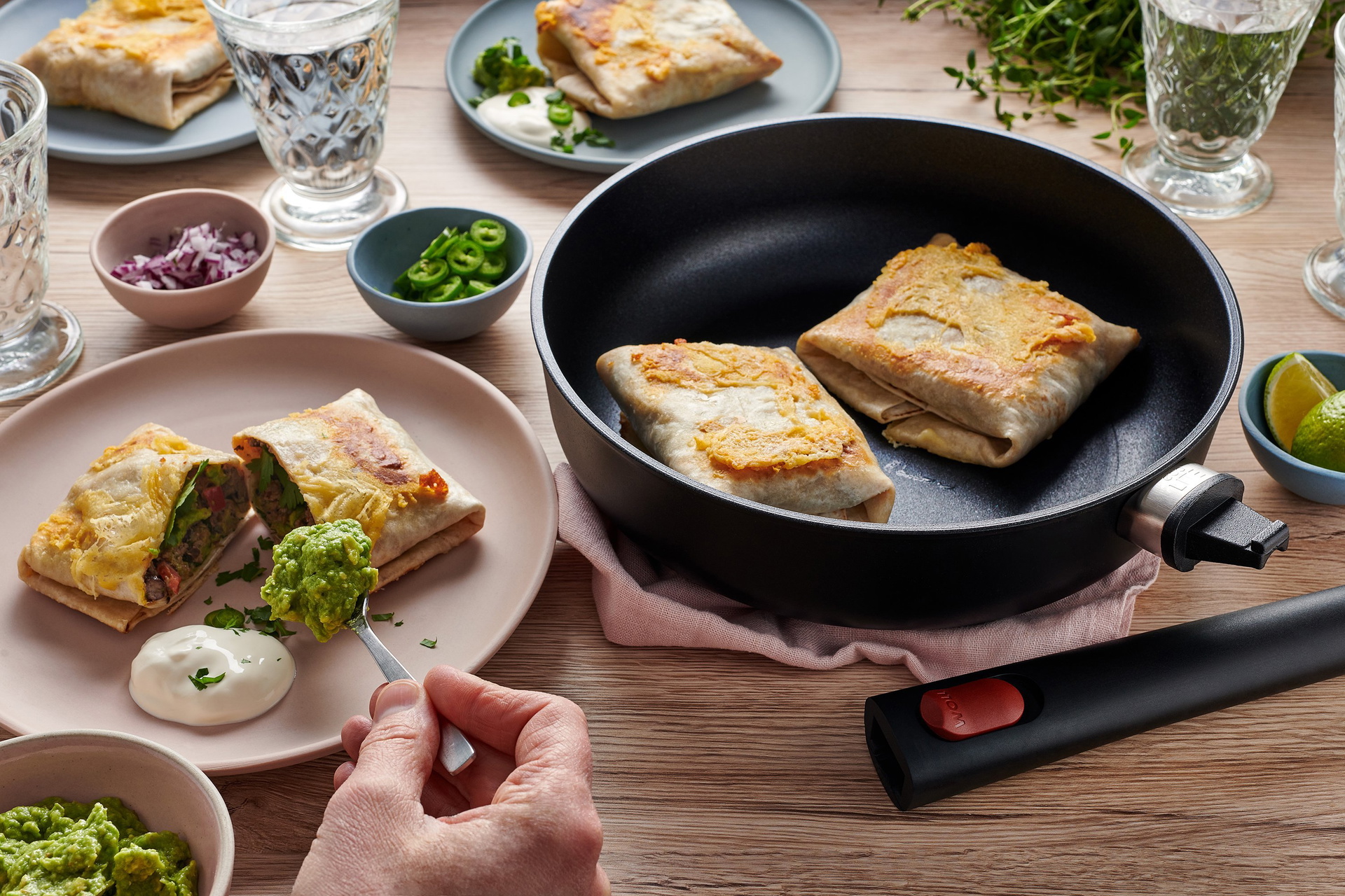 In einer runden Eco Lite QXR Pfanne mit abgenommenem Stiel befinden sich frisch angebratene Burritos. Ein Burrito ist in der Mitte geteilt und auf einem Teller angerichtet. Er wird mit zwei Dips serviert. Dahinter sind weitere gefüllte Teller und Gläser