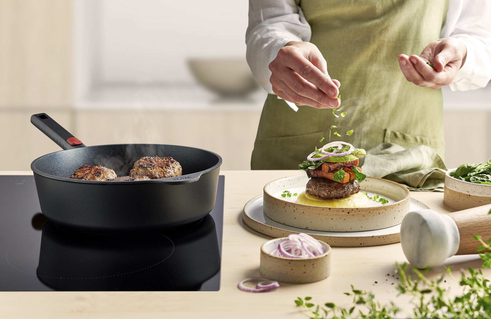 In einer runden Hochrandpfanne mit festem Holzstiel der Serie Eco Logic QXR liegt fertig angebrachtes Salisbury-Steak. Daneben steht ein Teller mit Kartoffelpüree, darauf ein Stück Salisbury-Steak, garniert mit Butternusskürbis-Chips, Spinat und Avocado.