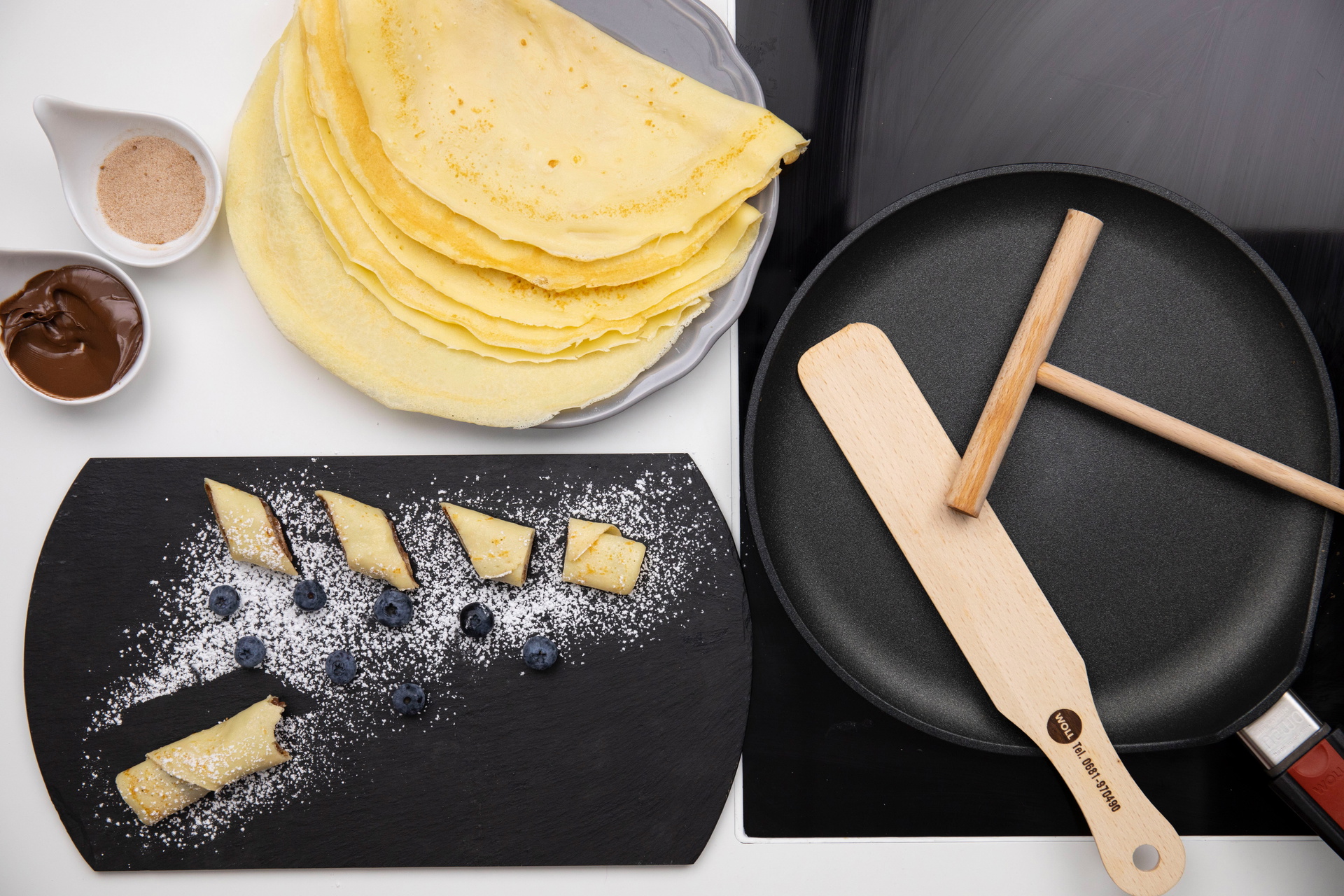 In einer runden Crêpepfanne der Serie Nowo mit abnehmbarem Stiel liegen ein Teigverteiler und ein Crêpeswender. Die fertig zubereiteten Crêpes liegen auf einem Teller daneben. Daneben stehen eine Schüssel mit Zimt und Zucker sowie eine weitere Schüssel mi