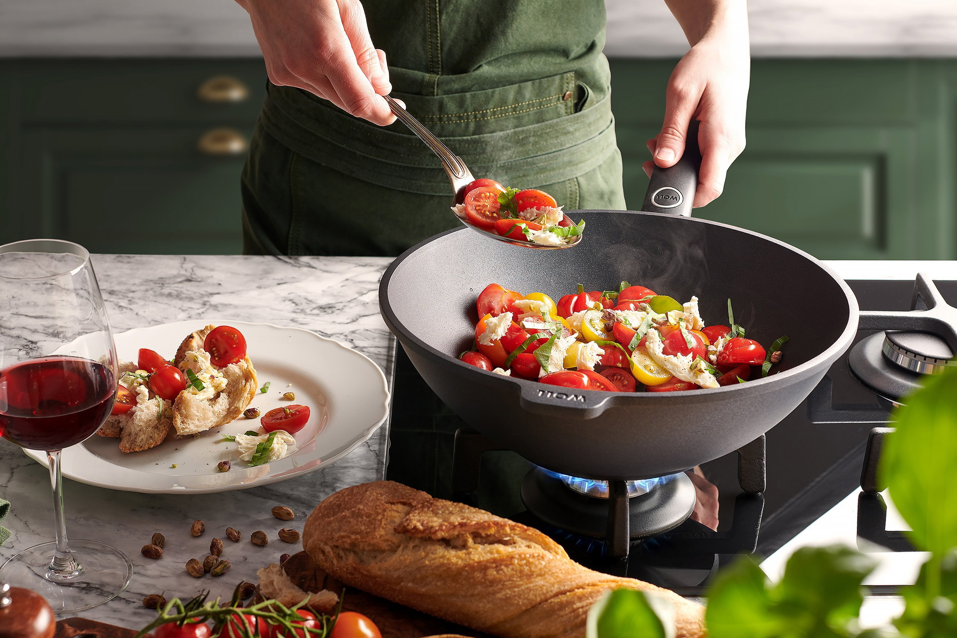 In einer runden Wok- und Rührpfanne wurde warmer Tomaten-Mozzarella-Salat zubereitet. Daneben stehen ein Teller, auf dem der Salat auf frischem Baguette angerichtet wurde, und ein Glas Rotwein. Drumherum liegen frisches Baguette und Cherrytomaten.