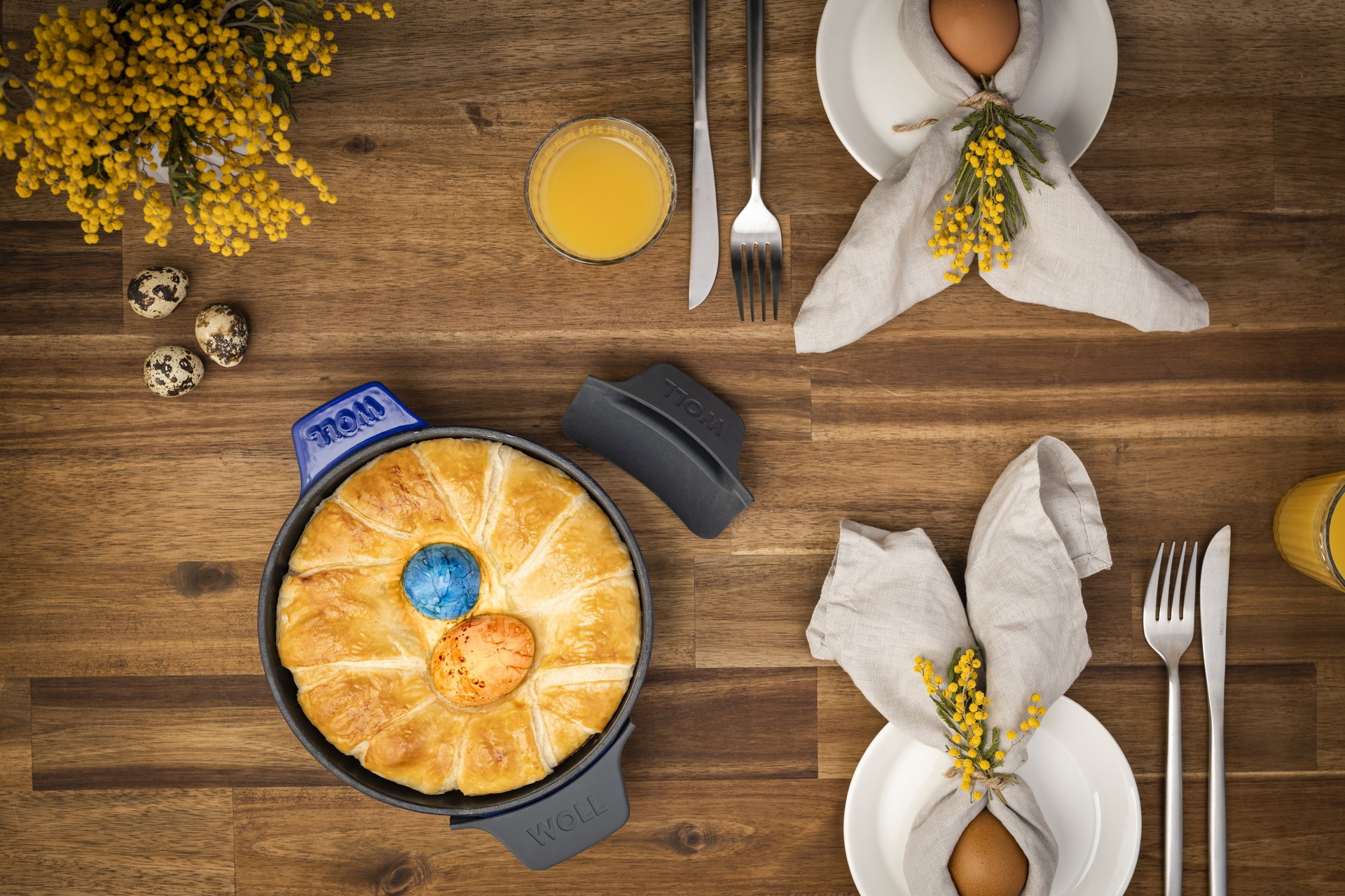 In einer runden Servierpfanne der Serie Iron befindet sich ein frisch gebackener Kuchen Ostereier. Zur Sicherheit wurden Silikongriffe um die Griffe gelegt. Ein Griff liegt daneben, damit die Farbe erkennbar ist. Daneben liegen gekochte Eier auf Tellern,