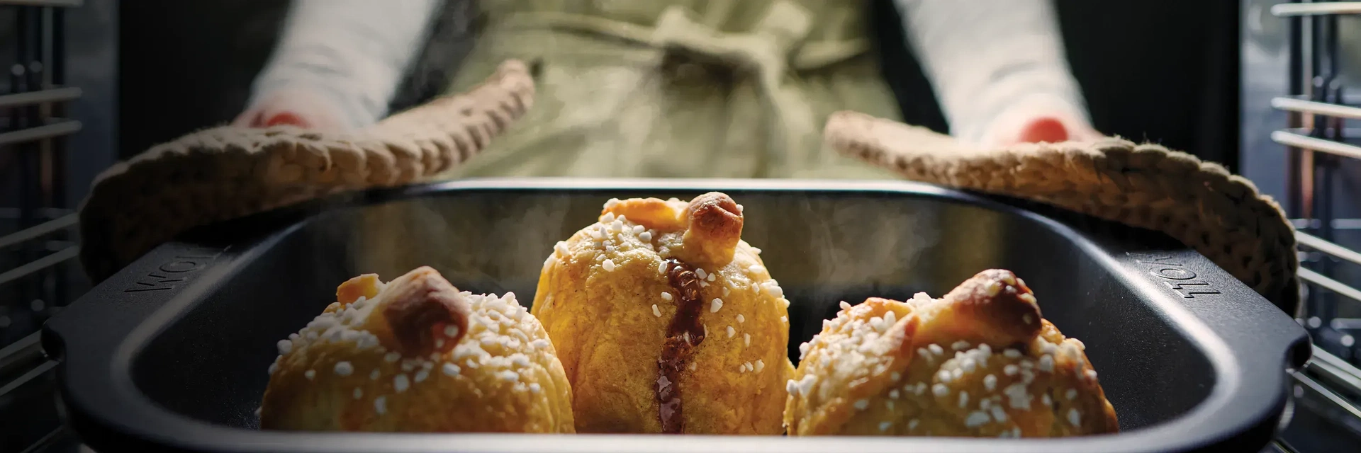 Eine heiße, viereckige Bratreine der Serie Roast it, beinhaltet ein dampfendes Gericht. Die Bratreine wird mit zwei Topflappen aus dem Backofen genommen.