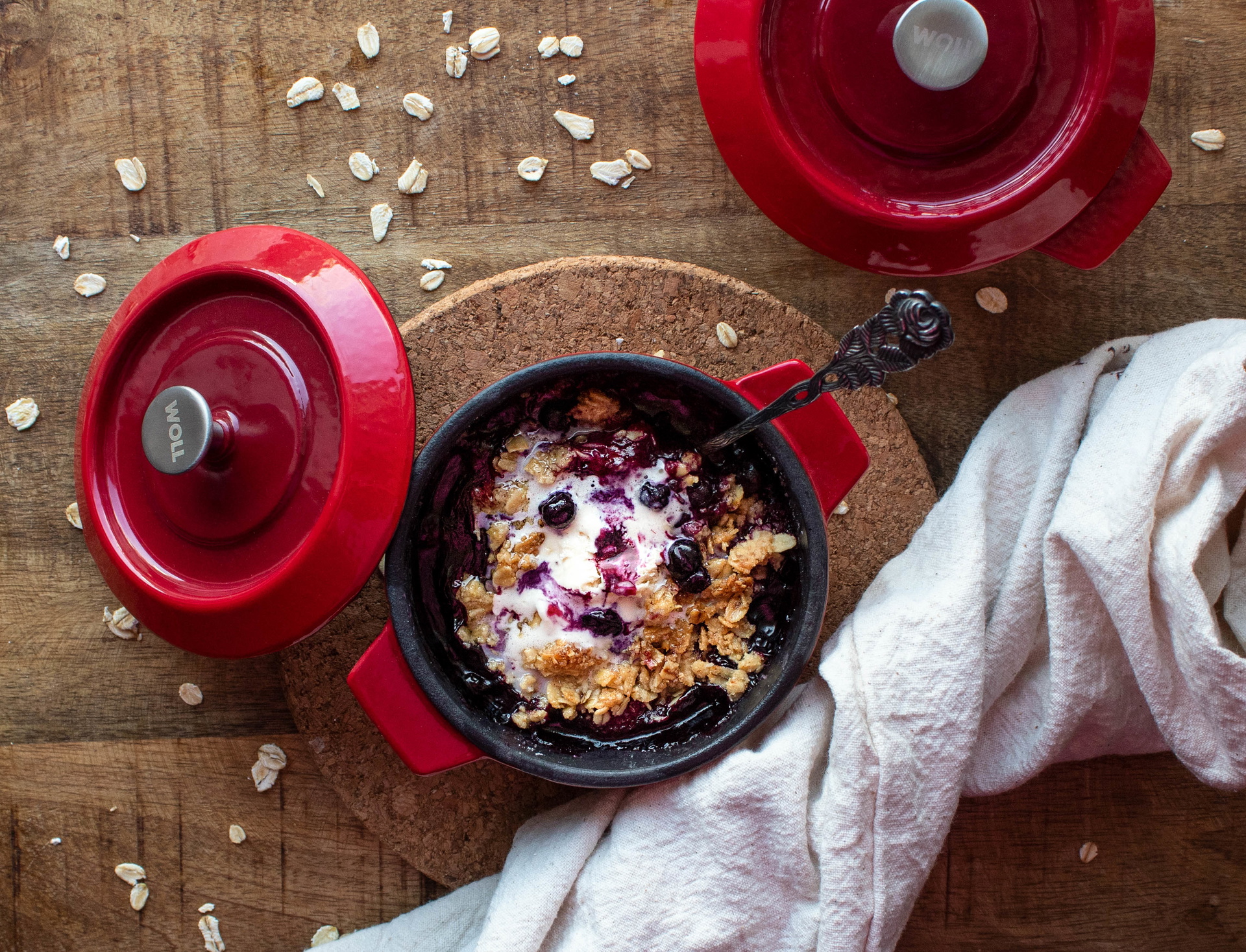 Auf einem Untersetzer steht eine Mini-Kasserolle der Serie „Iron”. Der Deckel wurde abgenommen und liegt schräg angelehnt daran. In der Kasserolle befindet sich frisch zubereitetes, warmes Beeren-Crumble. Daneben stehen eine weitere Kasserolle und ein Ges