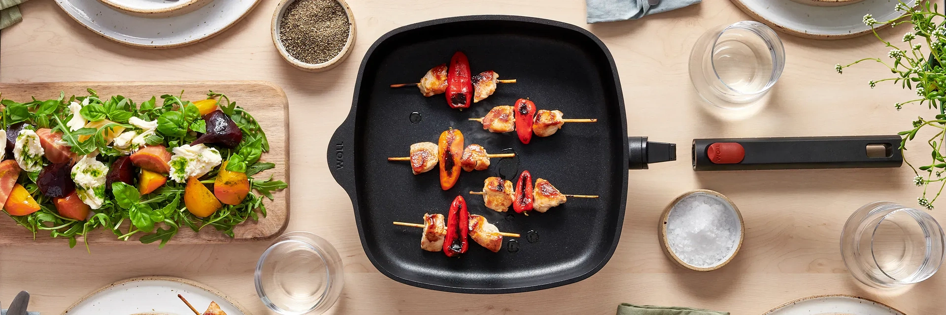 Auf dem Tisch steht eine viereckige Pfanne mit abgenommenem Stiel aus der Serie Eco Logic QXR, die frisch angebratene Fleischspieße enthält. Drumherum stehen Teller, Gläser, Gewürze und eine Pflanze.