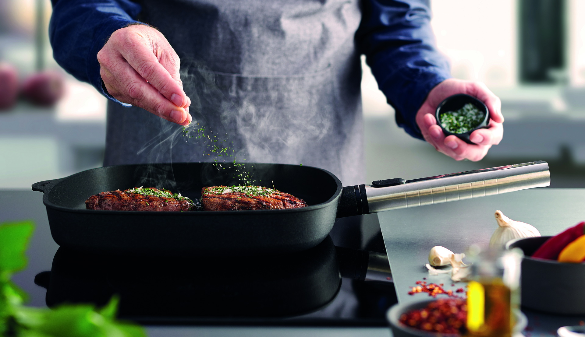Auf dem Herd steht die rechteckige, heiße Logic-Grillpfanne mit abnehmbarem Edelstahlstiel, in der saftig angebratenes Steak dampft. Es ist mit Kräutersalz bestreut. Daneben liegen frische Knoblauchzehen auf der Arbeitsplatte und in einem Behälter stehen