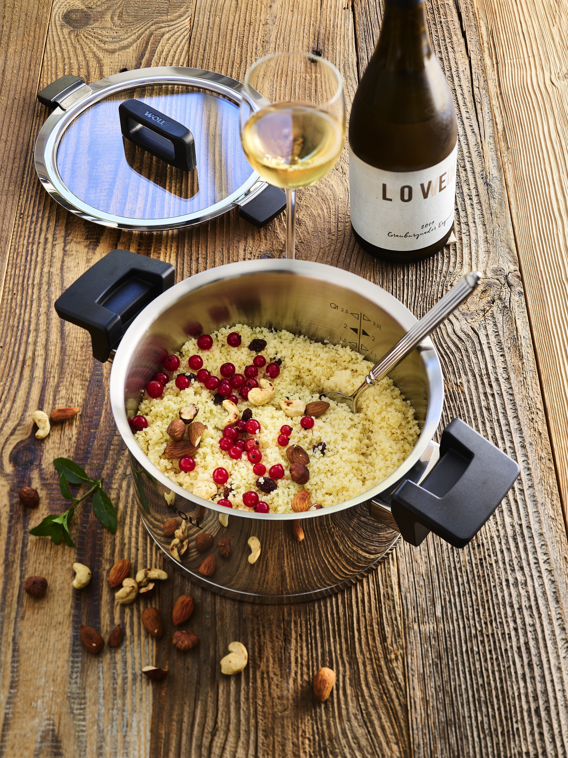 124NC_couscous_sweet_3543x4723_RGB In einem runden, heißen Concept-Topf befindet sich süßer Couscous mit Nüssen und Johannisbeeren.  Dahinter liegen der passende Glasdeckel, ein Weinglas und eine Weinflasche.