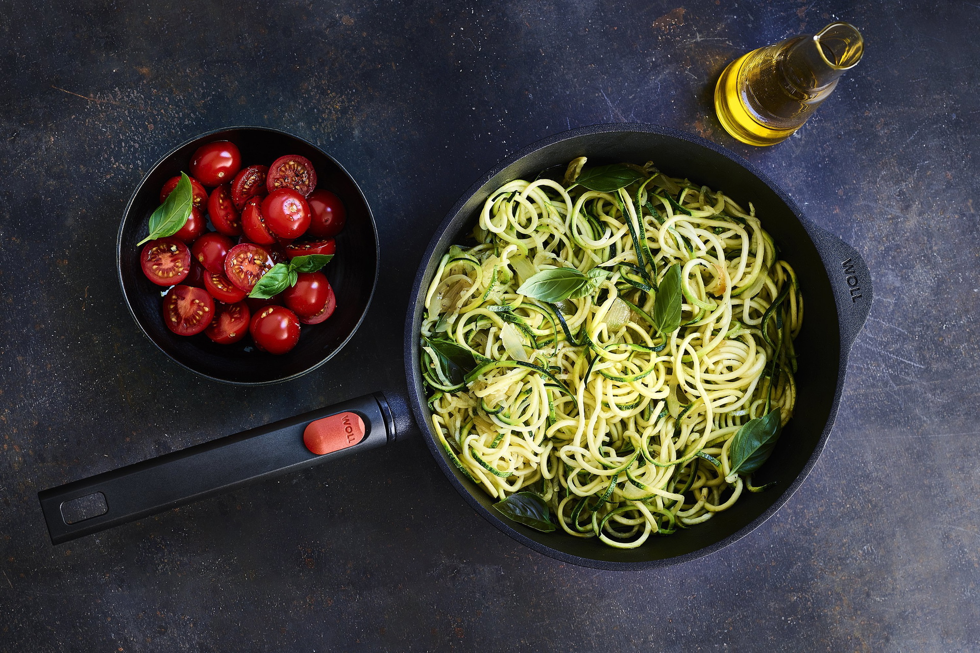 In einer runden, heißen Logic-Pfanne mit abnehmbarem Stiel befinden sich frisch zubereitete Zudeln, also Zucchinispaghetti. In einer daneben stehenden Schüssel befindet sich ein Kirschentomatensalat.