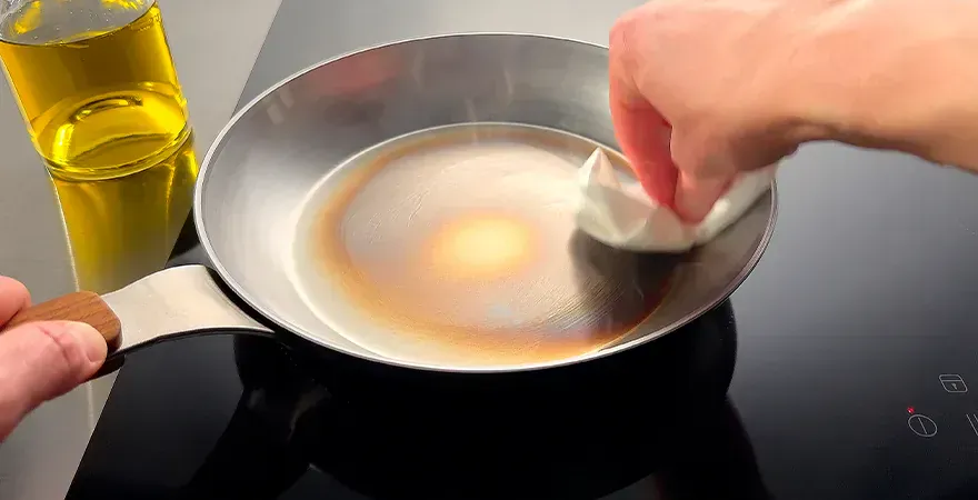 Auf dem Herd steht eine Carbon-Steel-Pfanne. In dieser wird mithilfe einer Küchenrolle das überschüssige Öl entfernt. Zur Fixierung wird die Pfanne am Griff festgehalten. Daneben steht eine Flasche mit neutralem Öl.
