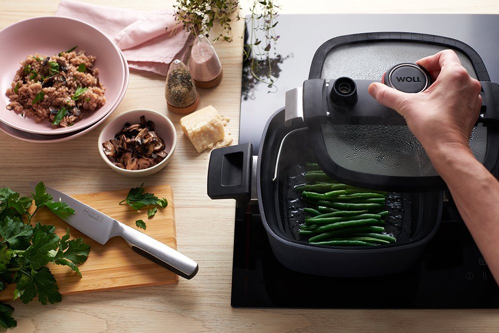 1022-2DALI_steamed_beans_1000px_RGB In einer heißen viereckigen Kasserolle der Serie Diamond Active Lite mit abgenommenem Glasdeckel werden in einem passenden Siebeinsatz Bohnen gedünstet. Auf der Arbeitsplatte links daneben liegt ein Schneidebrett, darauf frisch geschnittene Kräuter und ei
