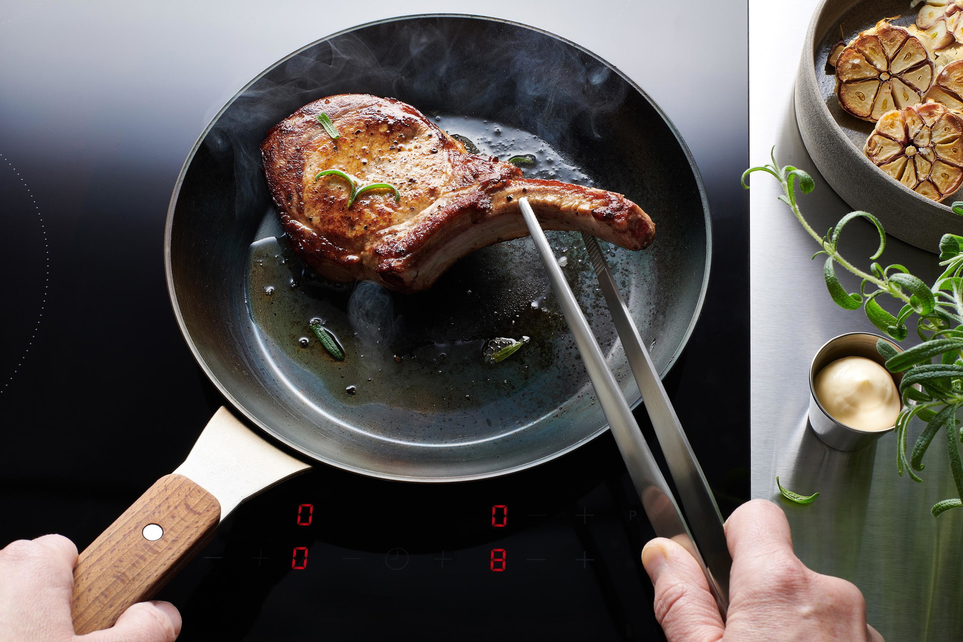 In einer Carbon-Steel-Pfanne mit eingebrannter Antihaftbeschichtung wird ein Stück Fleisch angebraten. Dieses wird mit einer Zange gewendet. Rechts daneben liegen angeröstete Knoblauchzehen und Kräuter.