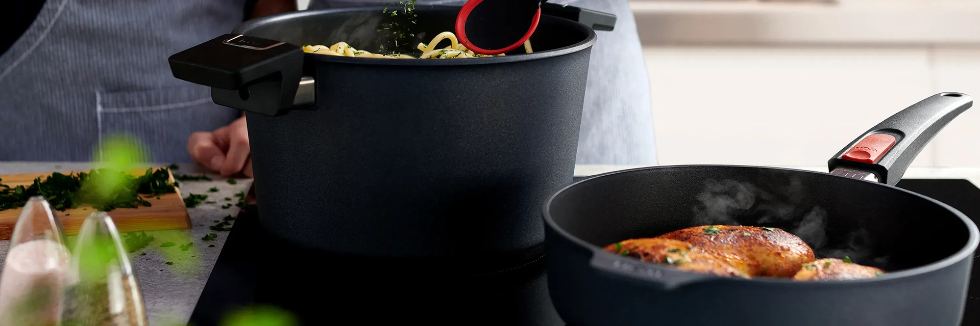 In einem Topf der Serie Diamond Lite werden frisch gekochte Nudeln mit einem Kochlöffel umgerührt und mit Kräutern garniert. Daneben steht eine runde, heiße Pfanne derselben Serie, in der sich dampfendes, goldbraun angebratenes Fleisch befindet. Auf der A
