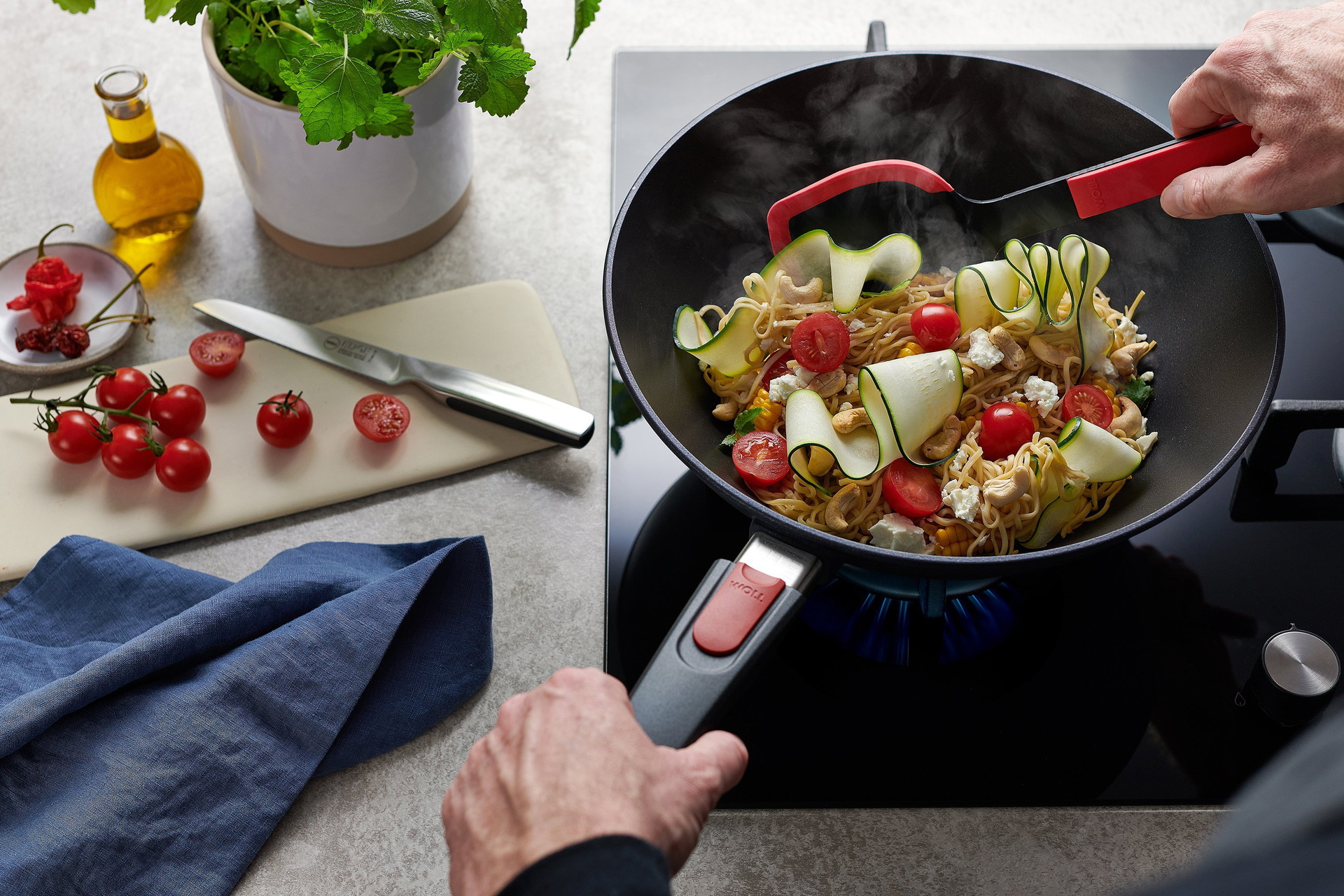 In einer runden Diamond Lite Wok- und Rührpfanne wird ein Nudelgericht zubereitet, das mit einer roten Cock it Pfannenschaufel umgerührt wird. 