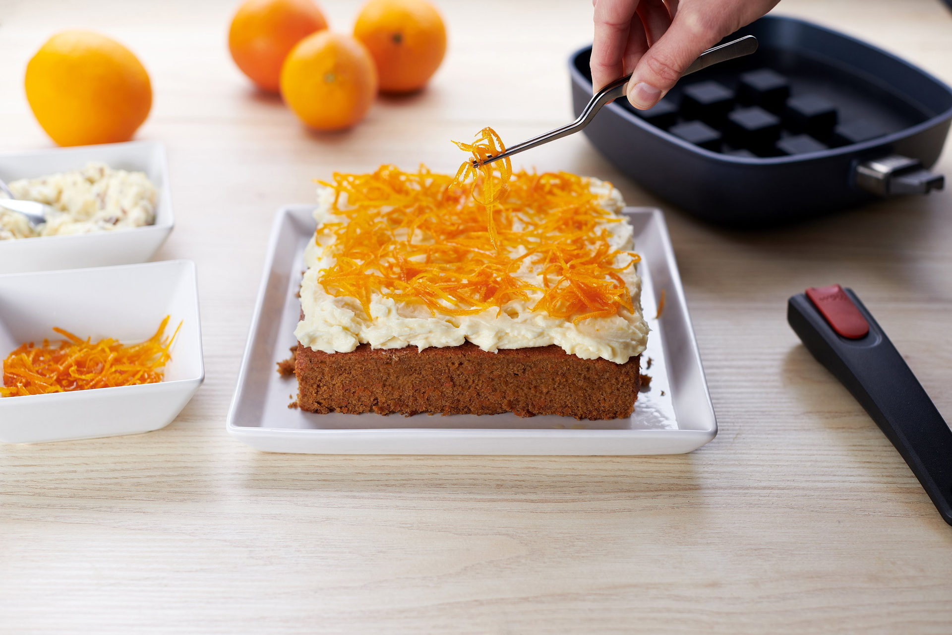 Ein frisch gebackener Kuchen mit cremigem Topping ist mit geriebener Orangenschale garniert. Im Hintergrund sind Orangen und eine Pfanne mit einem Silikon-Backeinsatz zu sehen. Daneben liegen der abgenommene Stiel der Pfanne sowie Schüsseln mit Creme und