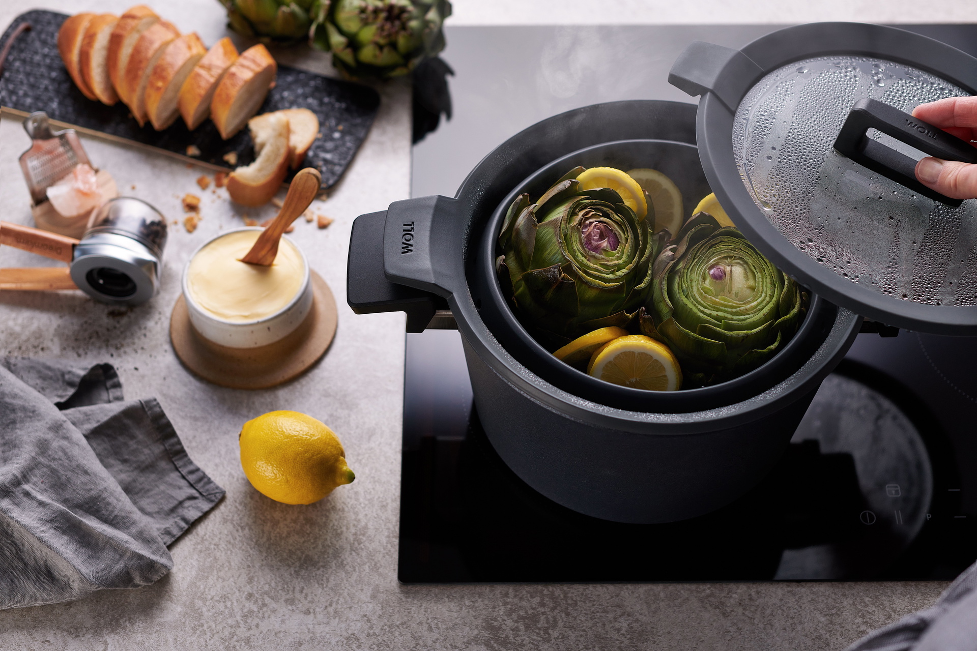 In einem runden Concept Plus Topf mit abgenommenem Glasdeckel und multifunktionalem Silikoneinsatz der Serie Steam it, werden Antischocken gedämpft. Daneben sind ein frisch geschnittenes Baguette, frische Butter, eine Mühle, eine Zitrone und ein Küchentuc