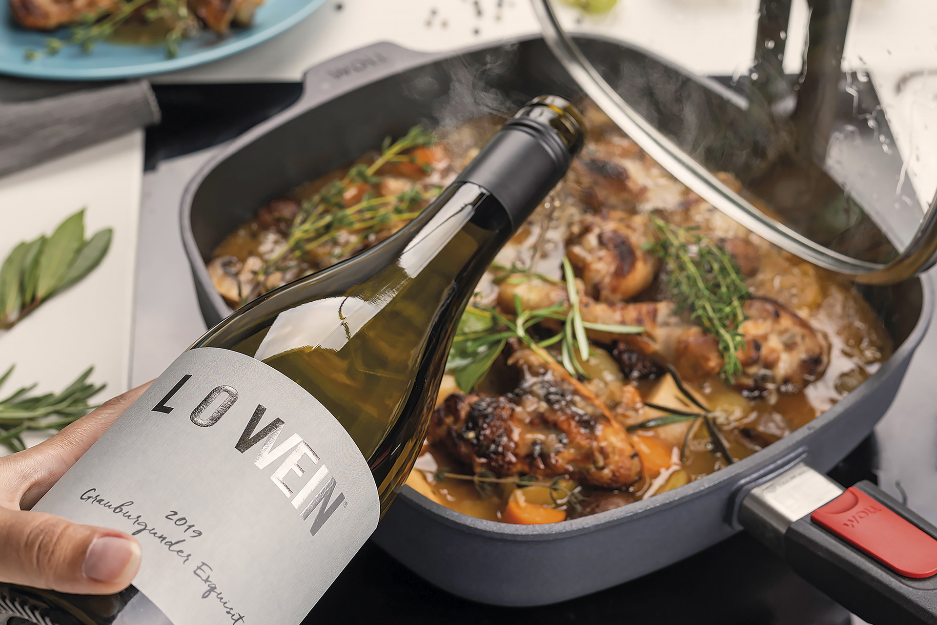 In einer viereckigen, heißen Diamond Lite Pfanne mit abnehmbarem Stiel wird frisches Coq au Vin zubereitet und mit Wein verfeinert. Der Glasdeckel wurde mit der verlängerten Seite der Smart-Lid-Serie an der Kante der Pfanne eingehängt. Auf der Arbeitsplat