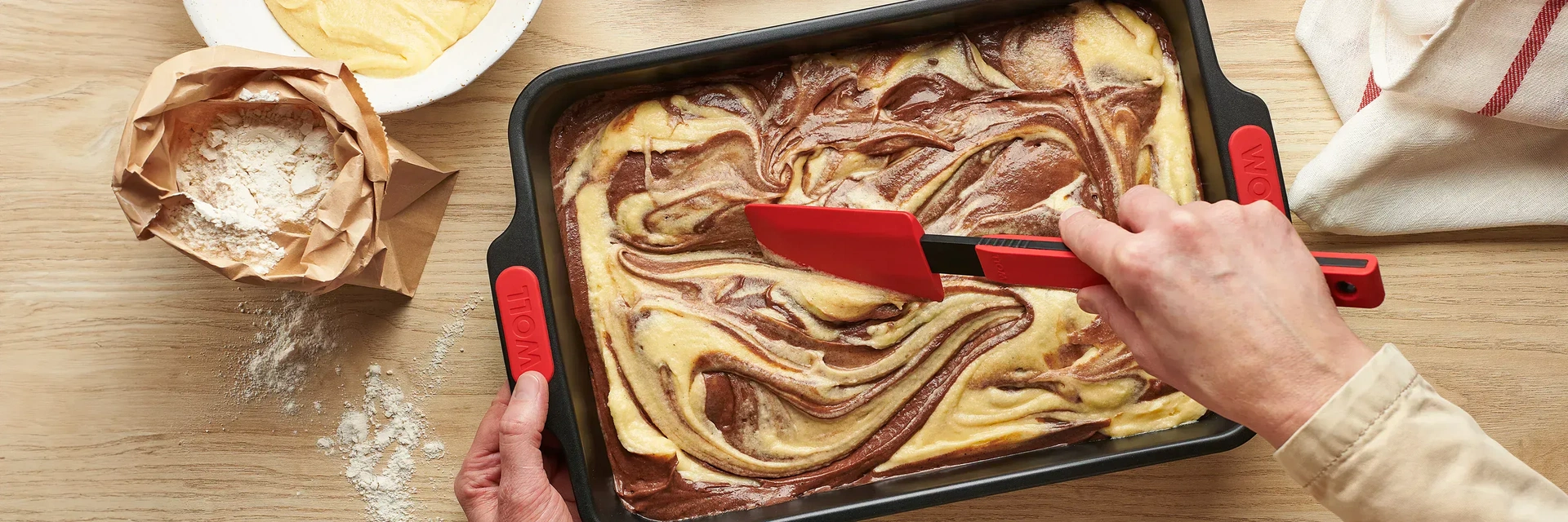 In der Backform der Serie Let’s Bake! befindet sich roher Kuchenteig, der mit einem Teigschaber der Serie Cook it” verteilt wird. Daneben sind eine offene Packung Mehl, ein Küchentuch sowie eine Schale mit Teig zu sehen.