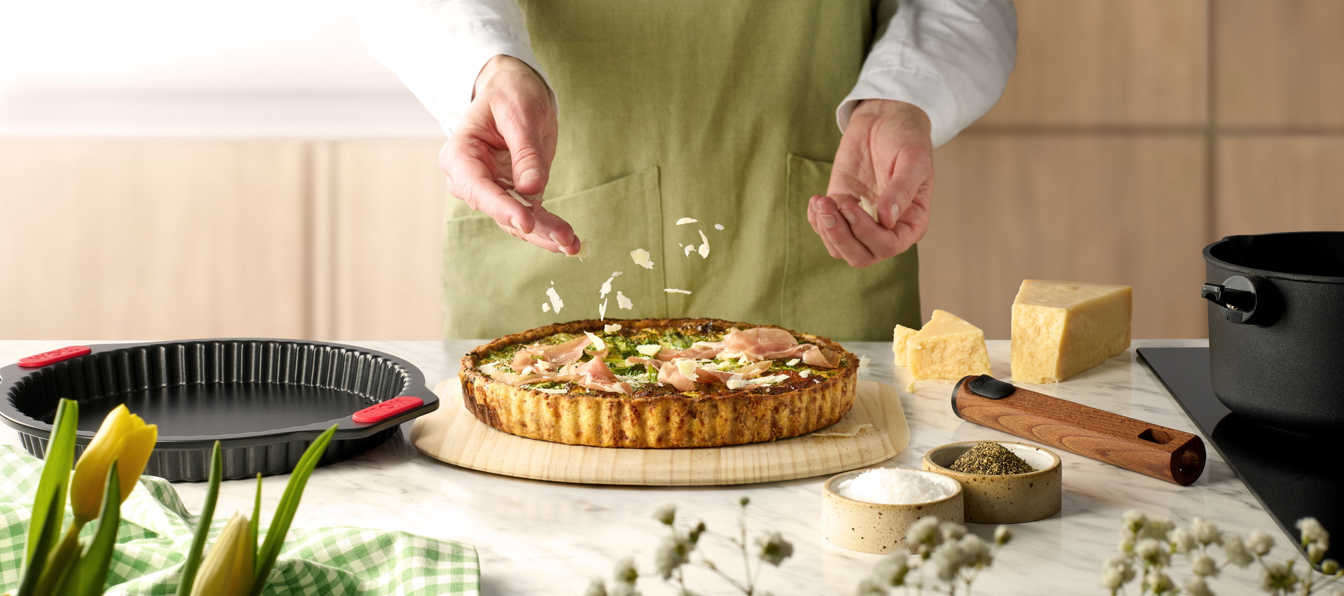 Auf einem Brett liegt eine frisch gebackene Frühlings-Quiche, die mit Schinken und Parmesan garniert ist. Sie wurde in einer Quiche- und Tarteform mit Hebeboden aus der Serie „Let's Bake!” gebacken. Drumherum liegen ein abgenommener Holzstiel, Parmesan un