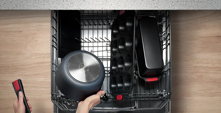 In der Spülmaschine befinden sich eine Muffinform und eine Kastenform der Serie Let's Bake!. Ebenfalls darin befinden sich ein Schneidebrett und ein Stieltopf, der gerade herausgenommen wird. Der Stiel des Topfes wurde abgenommen.