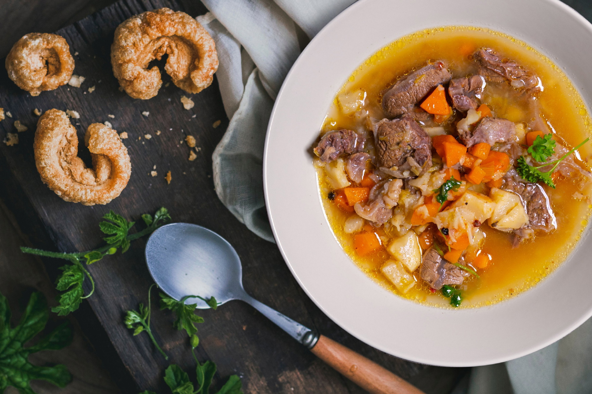 Auf einem Holzbrett steht ein Teller mit frisch zubereiteter Rindfleischsuppe. Daneben liegen ein Löffel und Brot.