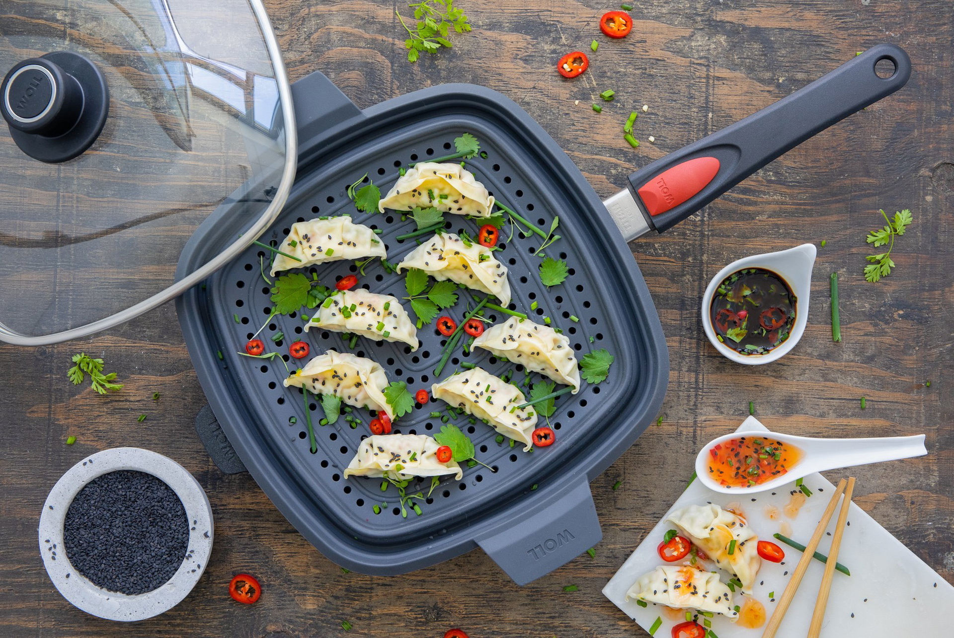 In einer viereckigen, heißen Titan-Best-Pfanne mit abnehmbarem Stiel wurden mithilfe eines multifunktionalen Silikoneinsatzes Dumplings gedämpft. Der Glasdeckel liegt auf der Kante der Pfanne. Daneben stehen Gefäße mit Öl und Soße. Darunter liegen die Dum